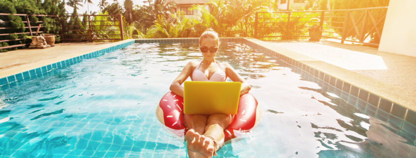 Frau in Badeanzug und mit Sonnenbrille sitzt im Schwimmring im Pool, genießt und benutzt Laptop zum Online-Shopping im Pool Shop
