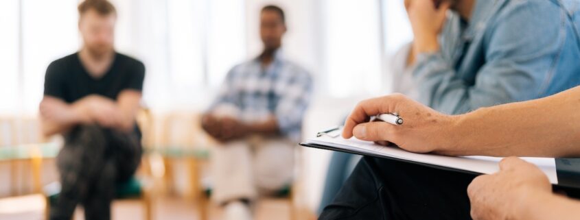 Personengruppe in Gesprächskreis mit Fokus auf Hand mit Stift und Klemmbrett im Vordergrund