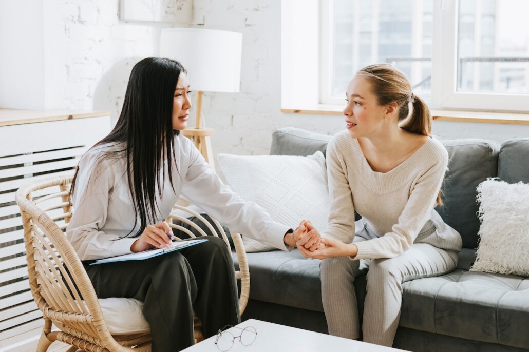 Zwei Frauen im Beratungsgespräch auf einem Sofa, eine hält die Hand der anderen einfühlsam
