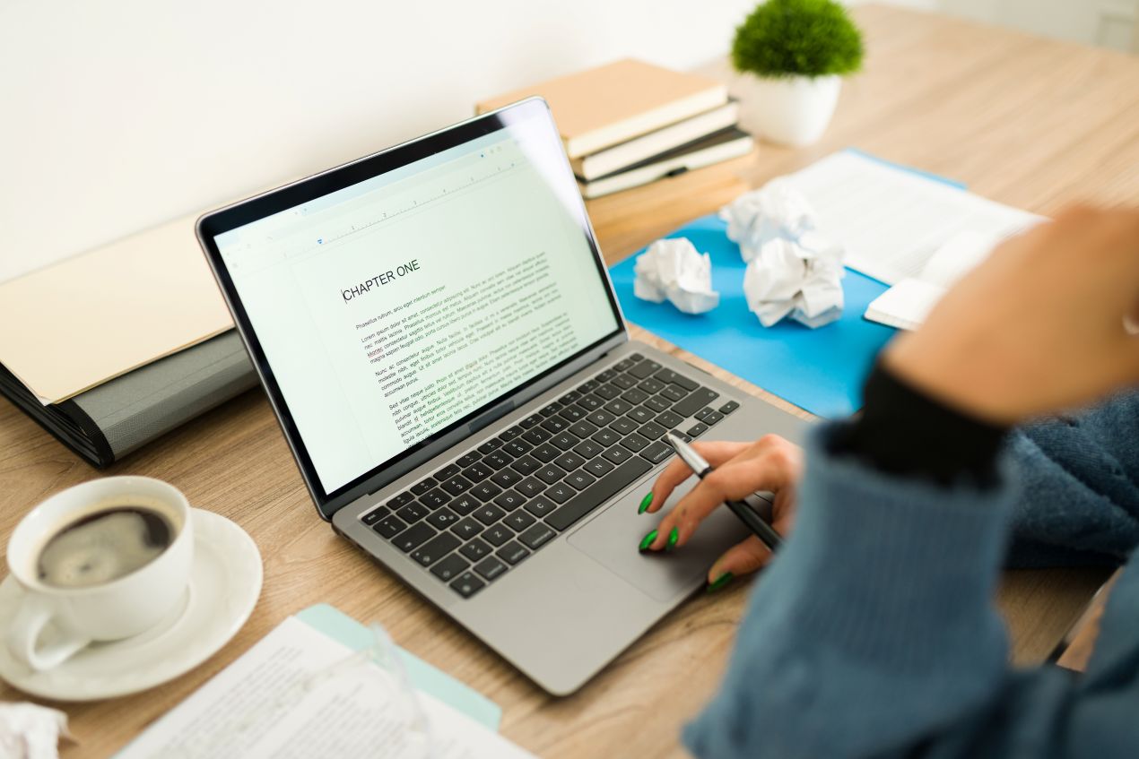 Laptop mit Kapitelanfang auf dem Bildschirm, Notizen, Kaffee und zerknülltes Papier – symbolisiert den intensiven Überarbeitungsprozess beim Buch Lektorat.